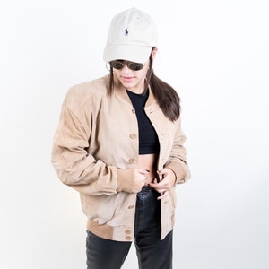 May include: A woman wearing a tan suede bomber jacket, black leather pants, and white sneakers. She is also wearing a white baseball cap with a small logo on the front and sunglasses.