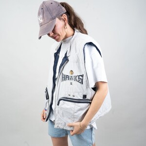 Op de afbeelding: Een vrouw draagt een wit T-shirt, een lichtgrijze vest met het woord "HAWKS" geborduurd op de voorkant, een blauwe denim short en een grijze baseballpet met de letters "NY" in wit.