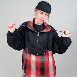 以下が含まれることがあります： 黒と赤のチェック柄のジャケットに、黒いジッパーと黒い帽子を合わせたコーディネート。ジャケットには、前面に大きなポケットが付いている。