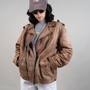 May include: A brown leather biker jacket with a zip fastening and multiple pockets. The jacket is worn over a grey t-shirt and white shorts. The person is wearing a brown baseball cap with a white "NY" logo.