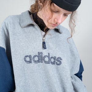 Puede incluir: Una sudadera de polar gris y azul con cierre de cremallera a la mitad y un logotipo de adidas azul marino en la parte delantera. La sudadera tiene un bolsillo con cremallera en la parte inferior derecha.