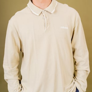 Op de afbeelding: Beige polo met lange mouwen, witte kraagafwerking en drie knopen. Het Adidas-logo is op de borst geborduurd. De shirt is gemaakt van een zacht, comfortabel materiaal.