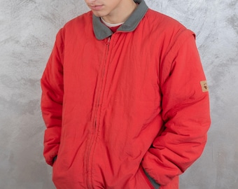 May include: A red zip-up jacket with a green collar and a brown patch on the right sleeve. The jacket is worn over a white t-shirt and beige trousers. The person is wearing a red and navy blue baseball cap with the text "CHICAGO BULLS" on the front.