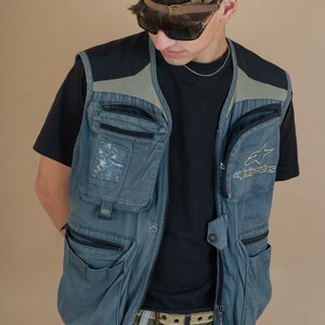 May include: A blue utility vest with multiple pockets and a black t-shirt underneath. The vest has a black mesh panel on the back and the Alpinestars logo on the right side. The person is wearing a brown baseball cap with blue star designs and sunglasses.