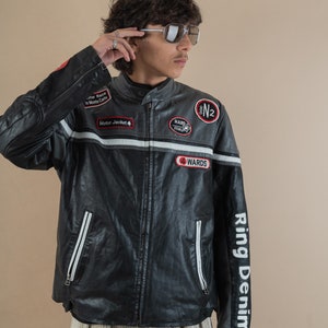 Puede incluir: Una chaqueta de cuero negra para moto con rayas blancas y varios parches. Los parches incluyen "Motor Jacket", "Hard Rock", "N2", "Wards" y "Star Race in Monte Carlo". La chaqueta tiene una cremallera y un logotipo blanco de "Ring Denim" en la manga derecha.