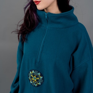 Könnte beinhalten: Türkisfarbener Fleece-Pullover mit hohem Kragen und Viertelreißverschluss. Der Pullover hat einen runden, gestickten Aufnäher mit gelbem und blauem Design. Das Model trägt silberne Creolen und hat lila Haare.