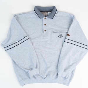 Puede incluir: Una camisa polo de manga larga gris con rayas negras en las mangas. La camisa tiene un cuello abotonado y un bolsillo con cremallera en el pecho izquierdo. La camisa tiene el texto "Classic Polo Sport" bordado en el bolsillo.