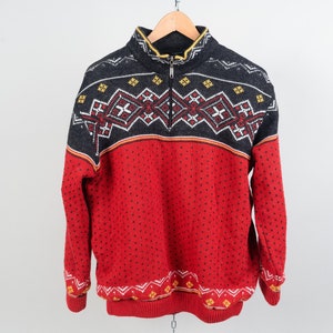 May include: A red, black and white patterned sweater with a zip up the front. The sweater has a high neck and long sleeves.