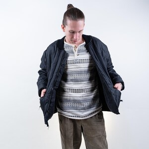 Könnte beinhalten: Eine schwarze gesteppte Bomberjacke mit Reißverschluss und zwei Taschen. Die Jacke wird über einem gestreiften Langarmshirt und einer braunen Hose getragen.
