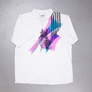 Op de afbeelding: Wit poloshirt met korte mouwen en een kleurrijk abstract ontwerp met roze, blauwe en zwarte strepen. Het shirt heeft een kraag en een knoopsluiting. Het woord "BOSS" is zichtbaar op het label.