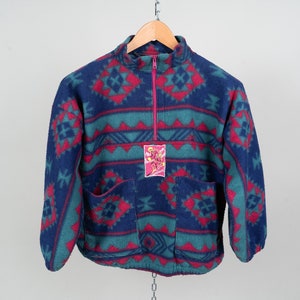 May include: A blue, green, and red fleece jacket with a geometric pattern. The jacket has a half-zip closure and two pockets. A pink and white label with the text "Sun Ice" is attached to the front of the jacket.