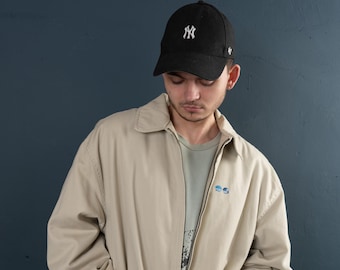 May include: A beige jacket with a zipper closure and two pockets. The jacket has a small embroidered logo on the left chest. The person is wearing a black baseball cap with the New York Yankees logo.