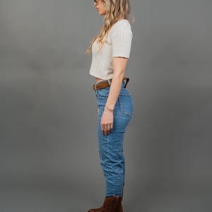 Peut inclure: Une femme porte un haut court blanc &agrave; manches courtes, un jean bleu, une ceinture marron et des bottes de cowboy marron. Elle porte &eacute;galement un bracelet et des lunettes. Le fond est gris uni.