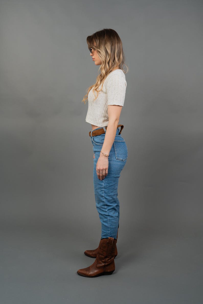 Peut inclure: Une femme porte un haut court blanc &agrave; manches courtes, un jean bleu, une ceinture marron et des bottes de cowboy marron. Elle porte &eacute;galement un bracelet et des lunettes. Le fond est gris uni.