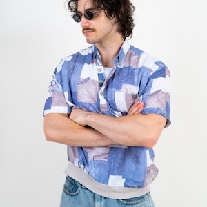 Könnte beinhalten: Ein kurzärmeliges Hemd mit Knopfleiste in einem blau-weiß-hellvioletten Patchwork-Muster mit Kettenmotiven. Das Hemd hat einen Kragen, eine halbe Knopfleiste und einen einfarbigen, hellen Saumbund und wird mit einer hellblauen Jeans kombiniert.