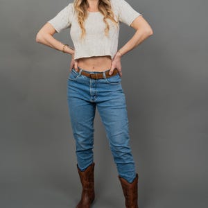 Peut inclure: Une femme porte un haut court cr&egrave;me &agrave; manches courtes, un jean bleu, une ceinture marron et des bottes de cowboy marron. Elle porte &eacute;galement des lunettes de soleil. Le fond est gris uni.