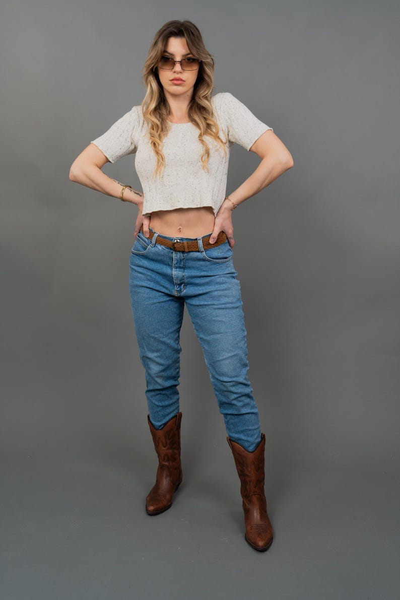 Peut inclure: Une femme porte un haut court cr&egrave;me &agrave; manches courtes, un jean bleu, une ceinture marron et des bottes de cowboy marron. Elle porte &eacute;galement des lunettes de soleil. Le fond est gris uni.