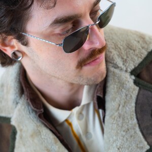 Op de afbeelding: Aviator zonnebril met een zilverkleurig montuur en donkere glazen. De persoon draagt een cr&egrave;mekleurig overhemd met kraag en een gele streep en een bruine en beige shearling jas.