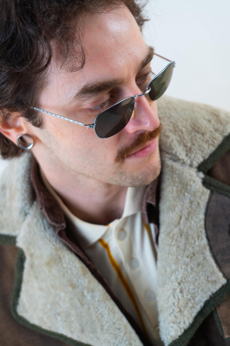 Op de afbeelding: Aviator zonnebril met een zilverkleurig montuur en donkere glazen. De persoon draagt een cr&egrave;mekleurig overhemd met kraag en een gele streep en een bruine en beige shearling jas.