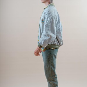 May include: A light blue bomber jacket with a green trim, worn over a patterned shirt and light green jeans. The person is wearing black and white sneakers.