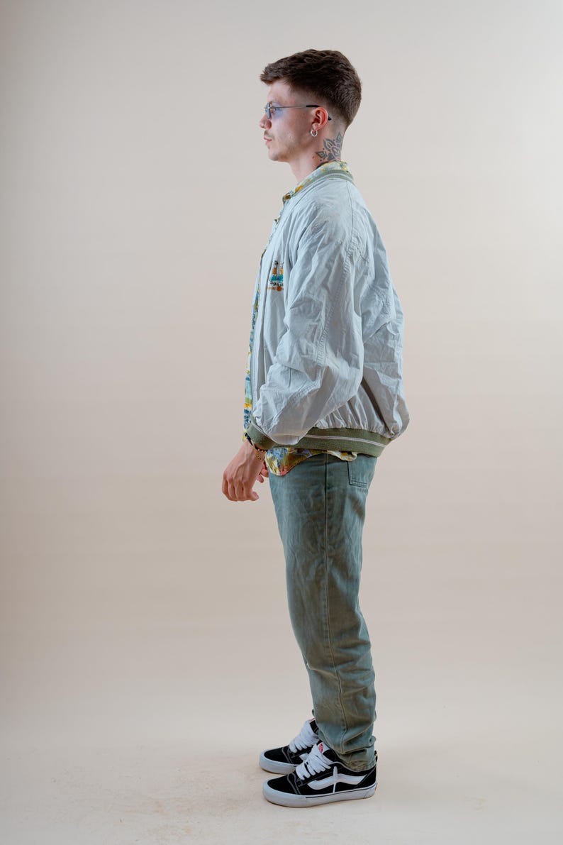 May include: A light blue bomber jacket with a green trim, worn over a patterned shirt and light green jeans. The person is wearing black and white sneakers.