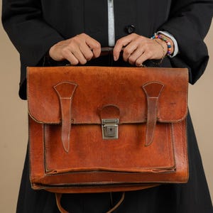 May include: A brown leather briefcase with a metal clasp and a strap. The briefcase is being held by a person wearing a black coat.