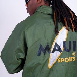 Könnte beinhalten: Eine olivgrüne Windjacke mit einer großen Grafik auf der Rückseite. Das Design zeigt eine weißes Surfbrett-Motiv mit gelber Umrandung, auf dem der schwarze Schriftzug MAUI und der gelbe Schriftzug SPORTS sowie ein Markenzeichen-Symbol zu sehen sind.