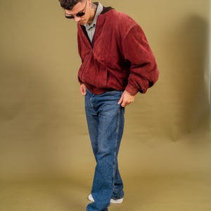 May include: A burgundy suede bomber jacket with a zip closure, paired with blue jeans and white sneakers. The outfit is completed with sunglasses. The style is casual and vintage.