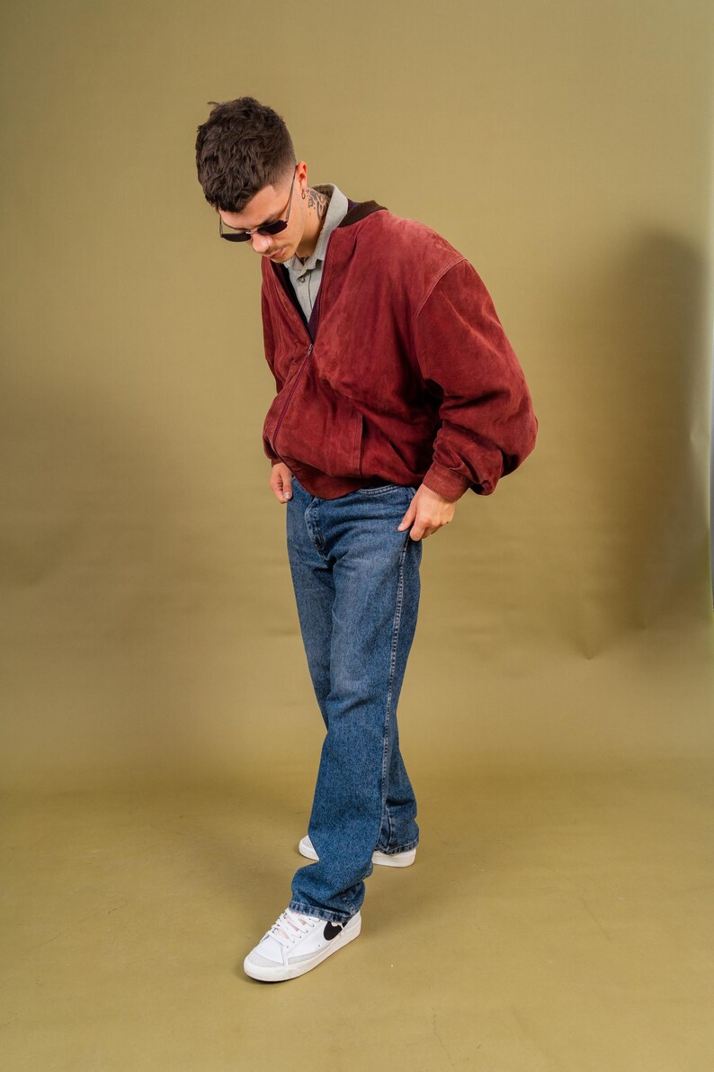 May include: A burgundy suede bomber jacket with a zip closure, paired with blue jeans and white sneakers. The outfit is completed with sunglasses. The style is casual and vintage.
