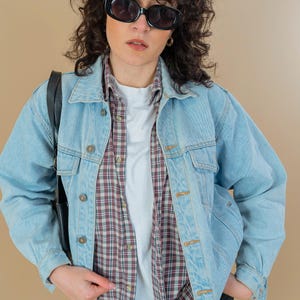 Könnte beinhalten: Eine Frau trägt eine hellblaue Jeansjacke über einem karierten Hemd und einem weißen T-Shirt. Sie trägt eine schwarze Sonnenbrille und eine schwarze Hose.