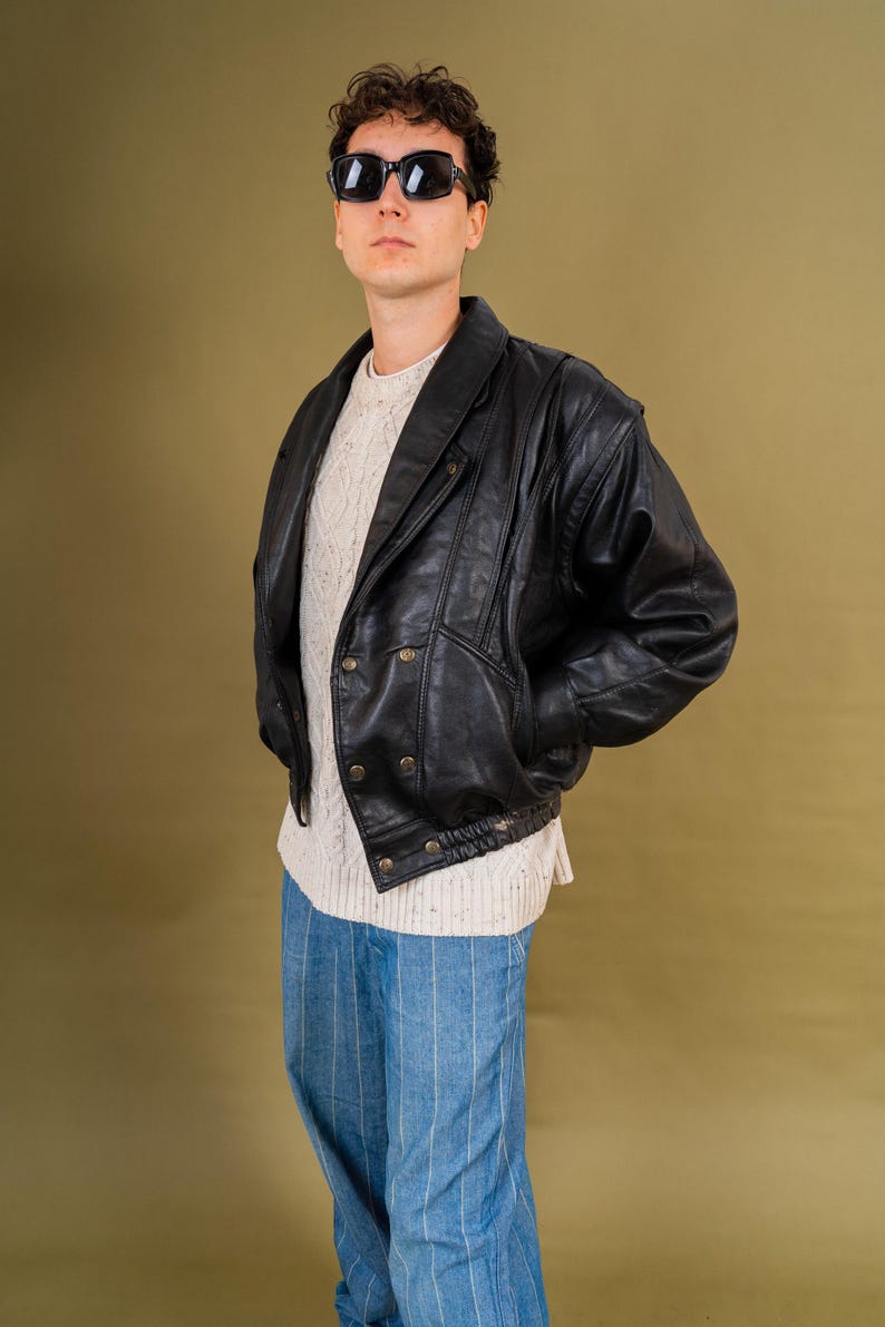 May include: A person wearing a black leather jacket with gold buttons, a white sweater, blue and white striped pants, and black sunglasses. The jacket has a wide collar and a ribbed hem. The outfit is vintage-inspired.