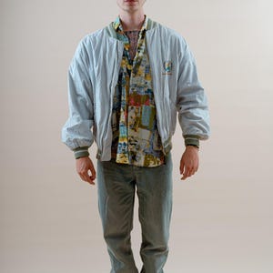May include: A light blue bomber jacket with a green trim, worn over a patterned button-up shirt and green pants. The outfit is completed with black and white sneakers. The jacket has a small emblem on the chest.