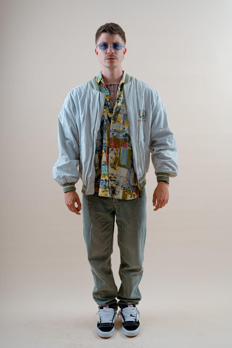 May include: A light blue bomber jacket with a green trim, worn over a patterned button-up shirt and green pants. The outfit is completed with black and white sneakers. The jacket has a small emblem on the chest.
