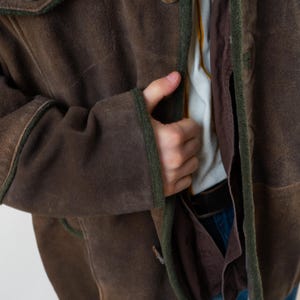 Op de afbeelding: Bruin su&egrave;de jasje met groene bies en houten knopen. Het jasje heeft een klassiek design met een getailleerde pasvorm. De mouwen zijn lang en de kraag is gekerfd. Het jasje wordt gedragen over een wit overhemd en een blauwe jeans.