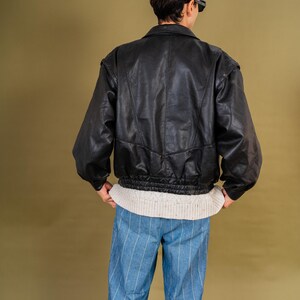 May include: A black leather jacket with a ribbed hem and shoulder pads. The jacket is worn over a cream-colored sweater and paired with blue and white striped pants. The person is wearing sunglasses.