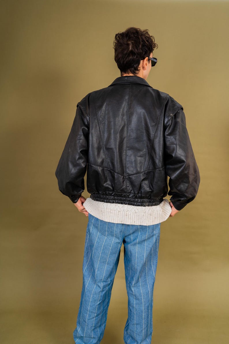 May include: A black leather jacket with a ribbed hem and shoulder pads. The jacket is worn over a cream-colored sweater and paired with blue and white striped pants. The person is wearing sunglasses.