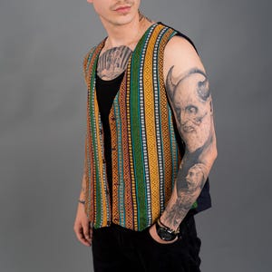 Op de afbeelding: Een patroonvest met verticale strepen in oranje, groen en blauw. Het vest wordt gedragen over een zwarte top en gecombineerd met een zwarte broek. De afbeelding is tegen een grijze achtergrond.