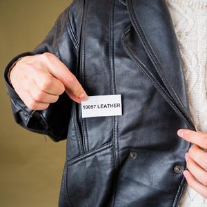 May include: Black leather jacket with a notched collar and button details. The jacket has a pocket and a tag that reads "10057 LEATHER". A cream-colored sweater is visible underneath the jacket.
