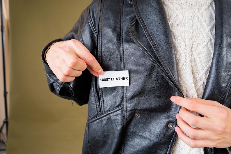 May include: Black leather jacket with a notched collar and button details. The jacket has a pocket and a tag that reads "10057 LEATHER". A cream-colored sweater is visible underneath the jacket.