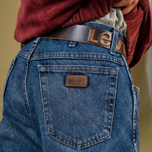 May include: Blue denim Wrangler jeans with a brown leather belt that reads "Lev" in embossed letters. The jeans have a brown leather patch with the Wrangler logo on the back pocket.
