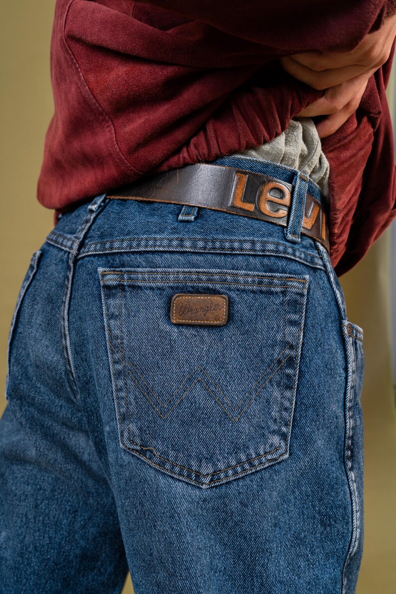 May include: Blue denim Wrangler jeans with a brown leather belt that reads "Lev" in embossed letters. The jeans have a brown leather patch with the Wrangler logo on the back pocket.