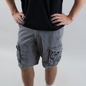 Könnte beinhalten: Graue Cargo-Shorts mit mehreren Taschen und einer Reißverschlusstasche am rechten Bein. Die Shorts werden von einer Person getragen, die ein schwarz-weiß gestreiftes Shirt und weiße Turnschuhe trägt.