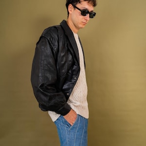 May include: A black leather jacket with a wide collar and a ribbed hem. The jacket is worn over a cream-colored sweater and paired with blue striped pants. The person is wearing black sunglasses.