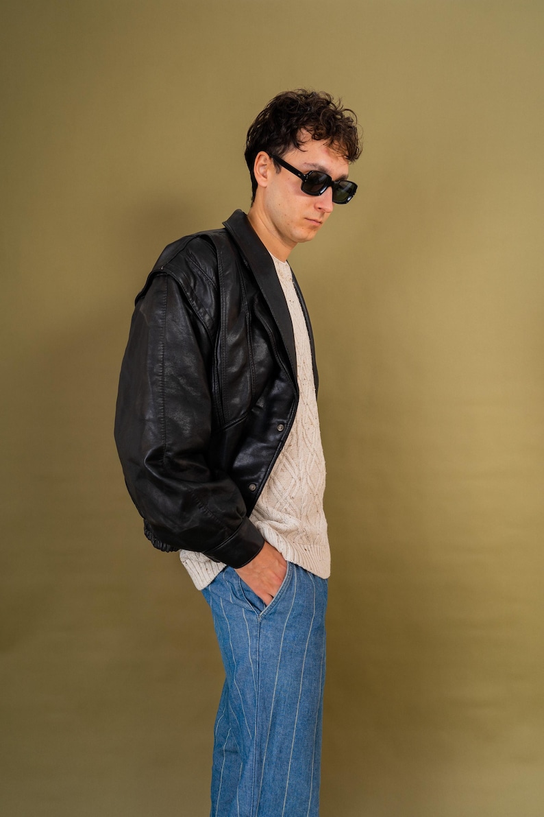 May include: A black leather jacket with a wide collar and a ribbed hem. The jacket is worn over a cream-colored sweater and paired with blue striped pants. The person is wearing black sunglasses.
