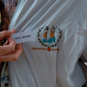 May include: A light gray jacket with a zipper closure and embroidered emblem. The emblem features two figures, a wreath, and the words "Adventure Repo". The tag reads "10217 JACKET".