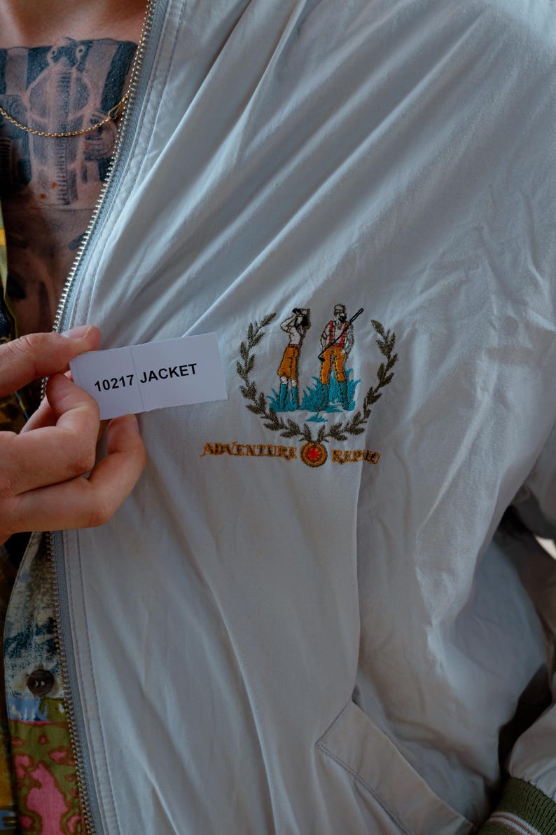 May include: A light gray jacket with a zipper closure and embroidered emblem. The emblem features two figures, a wreath, and the words "Adventure Repo". The tag reads "10217 JACKET".