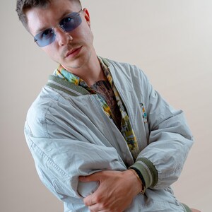 May include: A light gray bomber jacket with green trim on the cuffs and collar. The jacket is worn over a patterned shirt and paired with blue-tinted sunglasses. A gold bracelet is visible on the wrist.