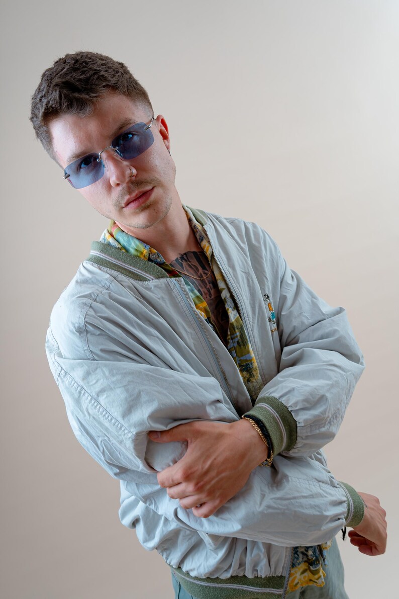 May include: A light gray bomber jacket with green trim on the cuffs and collar. The jacket is worn over a patterned shirt and paired with blue-tinted sunglasses. A gold bracelet is visible on the wrist.