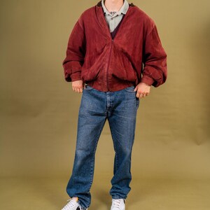 May include: A person wearing a burgundy suede bomber jacket with a zip closure, over a light green collared shirt and dark purple sweater. The person is also wearing blue denim jeans and white sneakers.