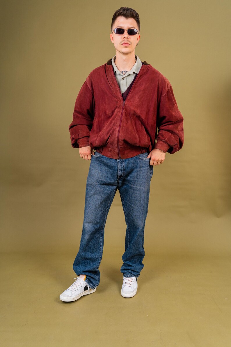 May include: A person wearing a burgundy suede bomber jacket with a zip closure, over a light green collared shirt and dark purple sweater. The person is also wearing blue denim jeans and white sneakers.